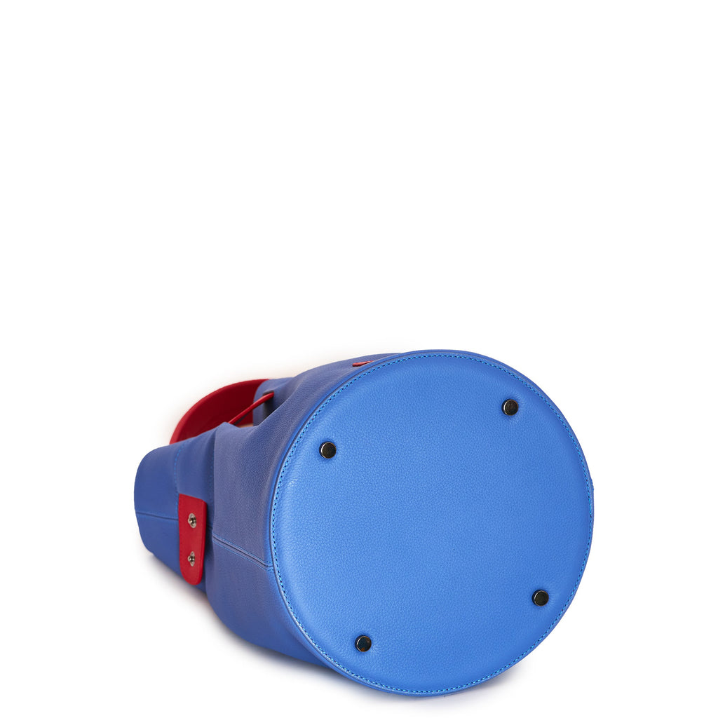 The product photo of a colorful fashion handbag showing a blue leather bucket bag with a red belt cinch and a red top handle. The bottom of the bag is round and has 4 gunmetal rivets. This is the bottom view of the Tomoli La Bourse leather bucket bag in Hot Iris. 