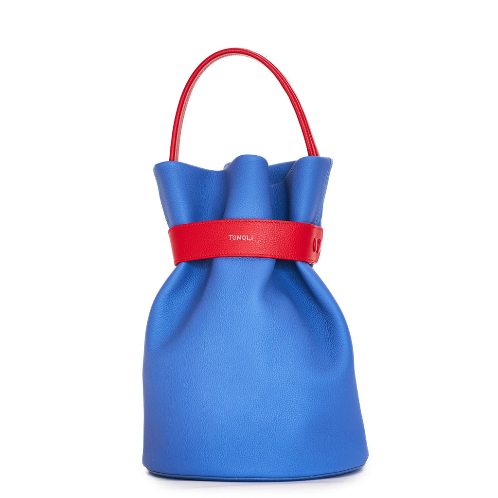 The product photo of a colorful fashion handbag showing a blue leather bucket bag with a red belt cinch and a red top handle. This is the front view of the Tomoli La Bourse leather bucket bag in Hot Iris.