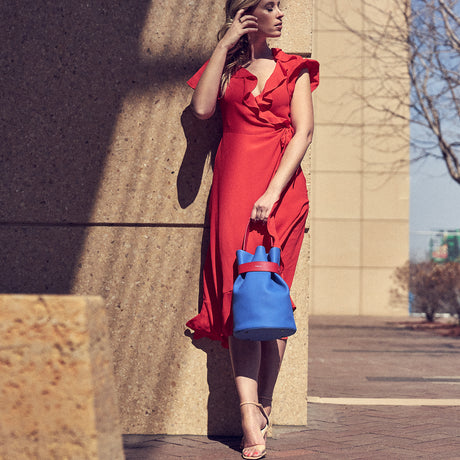 An outfit and style inspiration photo on how to wear a bucket bag. A woman wearing an orange summer dress is holding a blue leather bucket bag.  The handbag has a red belt cinch and a red handle. The woman's outfit is color-blocked. The bag is the Tomoli La Bourse leather bucket bag in Hot Iris.