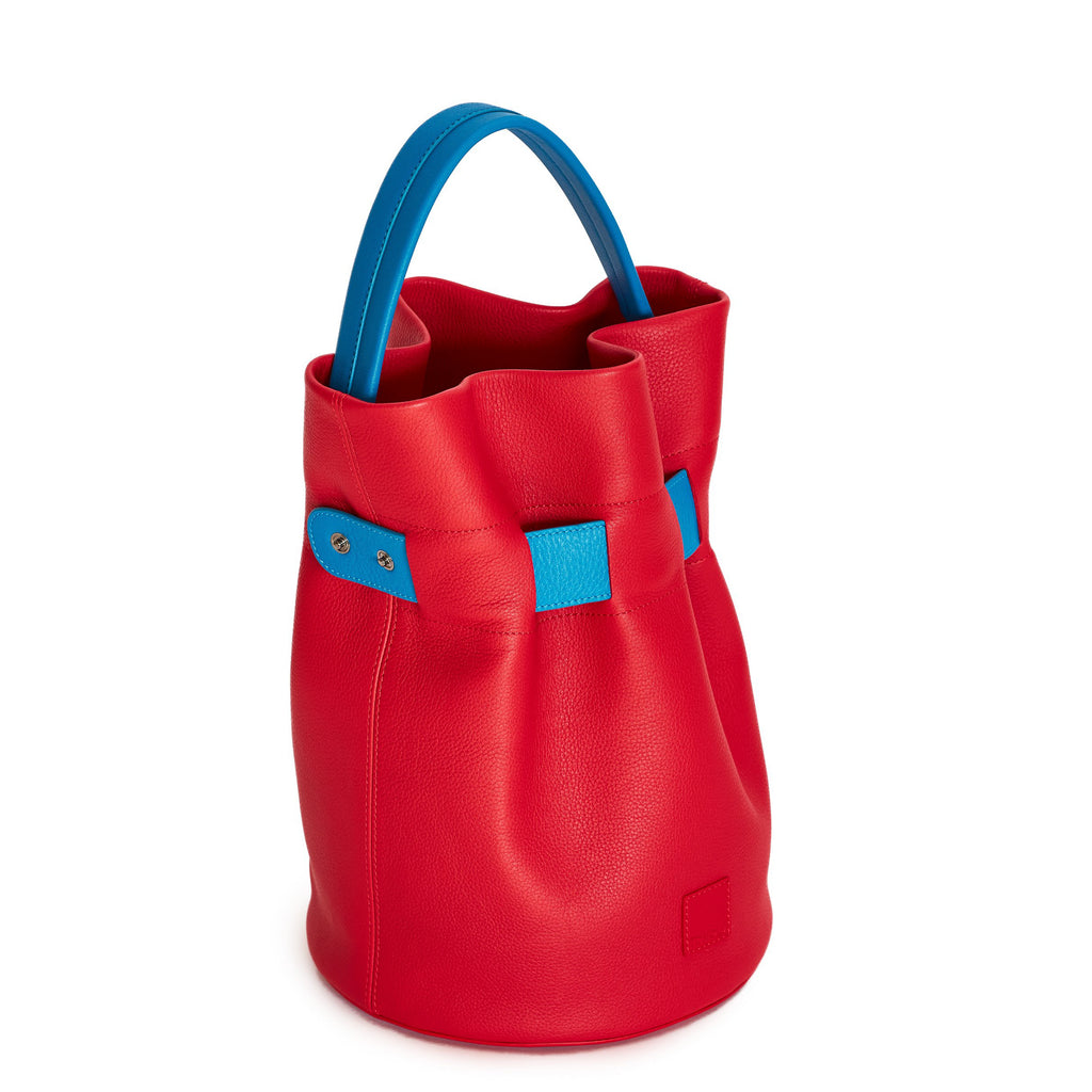 The product photo of a colorful fashion handbag showing a red leather bucket bag with a blue belt cinch and a blue top handle. The belt is woven into the bag and has 2 gunmetal snap buttons. This is the side view of the Tomoli La Bourse leather bucket bag in Sky Red.