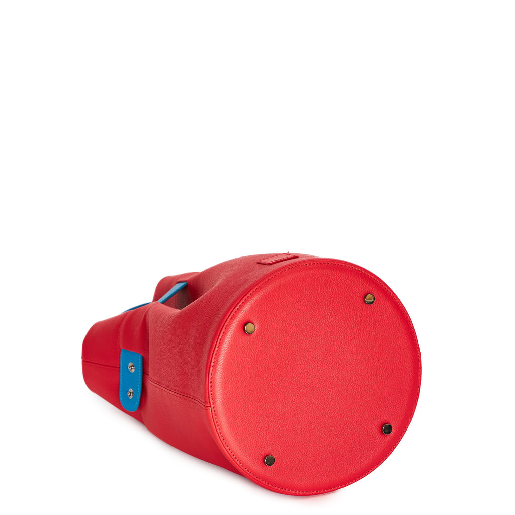The product photo of a colorful fashion handbag showing a red leather bucket bag with a blue belt cinch and a blue top handle. The bottom of the bag is round and has 4 gunmetal rivets. This is the bottom view of the Tomoli La Bourse leather bucket bag in Sky Red.