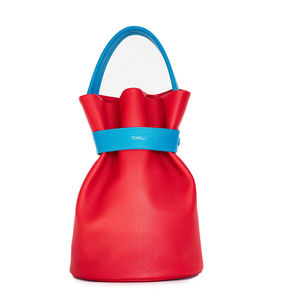 The product photo of a colorful fashion handbag showing a red leather bucket bag with a blue belt cinch and a blue top handle. This is the front view of the Tomoli La Bourse leather bucket bag in Sky Red.