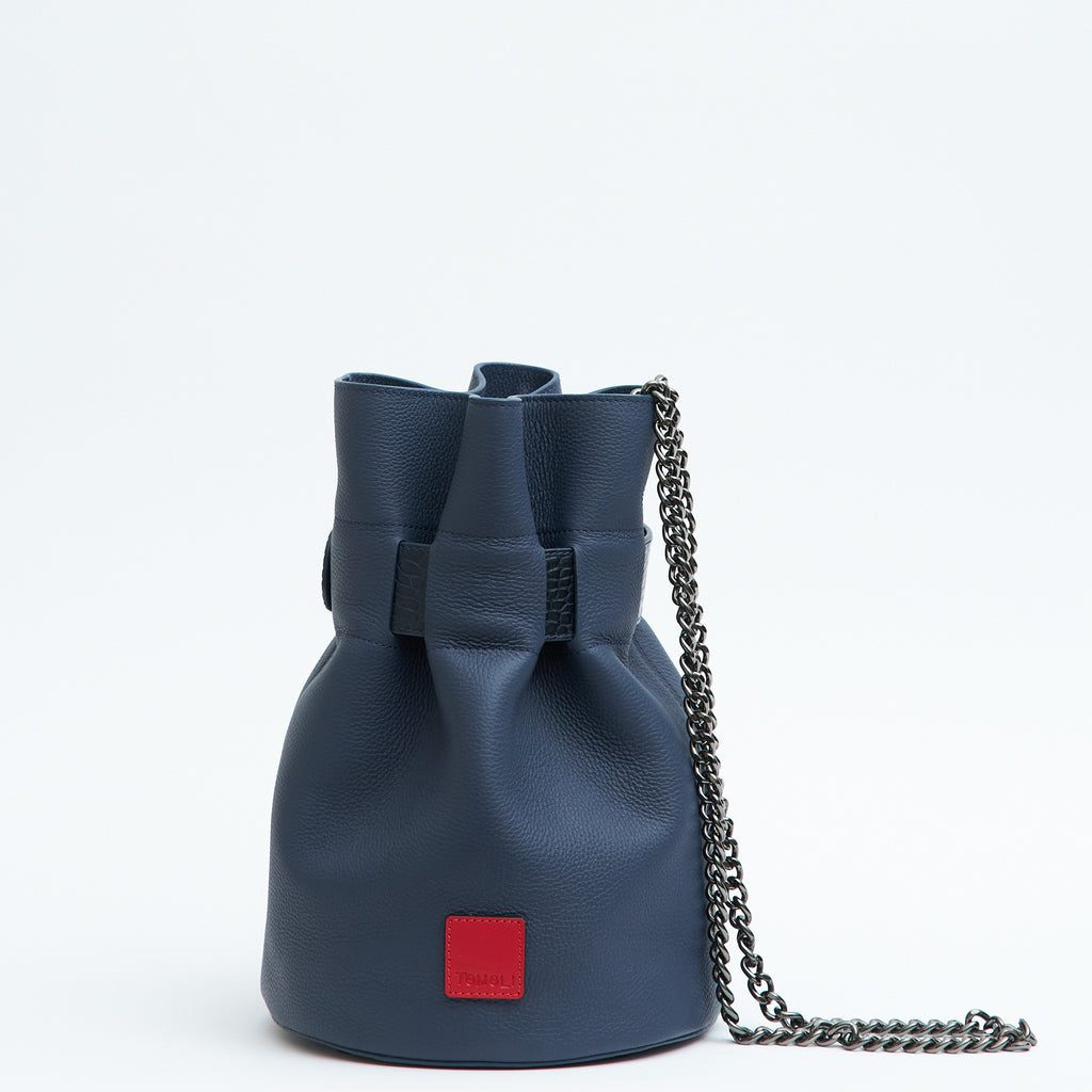 The product photo of a fashion handbag showing a dark blue leather bucket bag with a croc-embossed belt cinch and a chain strap. This is the back view of the Tomoli La Bourse Mini leather bucket bag in Denim.