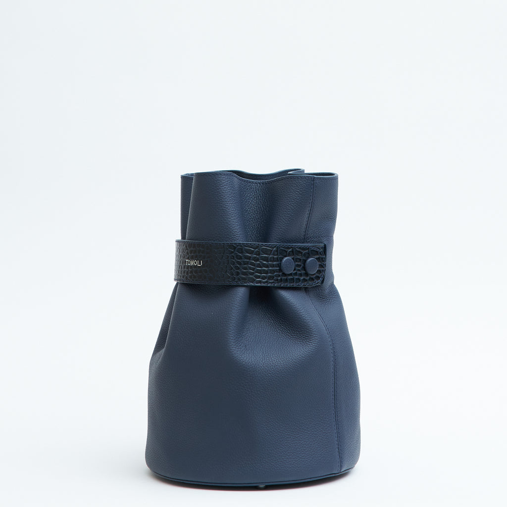 The product photo of a fashion handbag showing a dark blue leather bucket bag with a croc-embossed belt cinch. The belt has an adjustable snap closure on the side. This is the side view of the Tomoli La Bourse leather bucket bag in Denim.