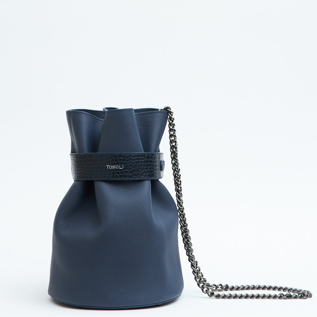 The product photo of a fashion handbag showing a dark blue leather bucket bag with a croc-embossed belt cinch and a chain strap. This is the front view of the Tomoli La Bourse Mini leather bucket bag in Denim.