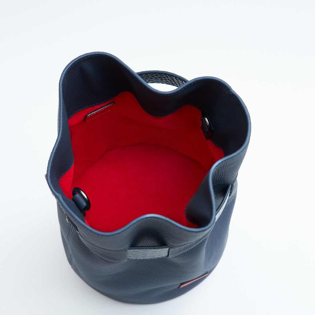 The product photo of a fashion handbag showing the interior of a dark blue leather bucket bag. The bag has a bright red lining made of alcantara microsuede material. This is the interior view of the Tomoli La Bourse Mini leather bucket bag in Denim.