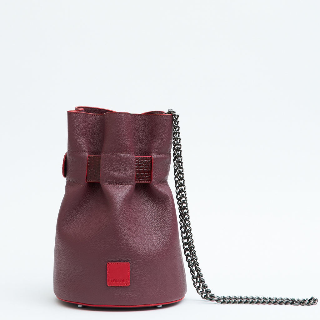 A product photo showing the back view of a dark red leather bucket bag. The bag has a croc-embossed belt cinch that is woven across the top. The TOMOLI logo is debossed on a small red square at the bottom of the bag. There is a chain strap in gunmetal color.