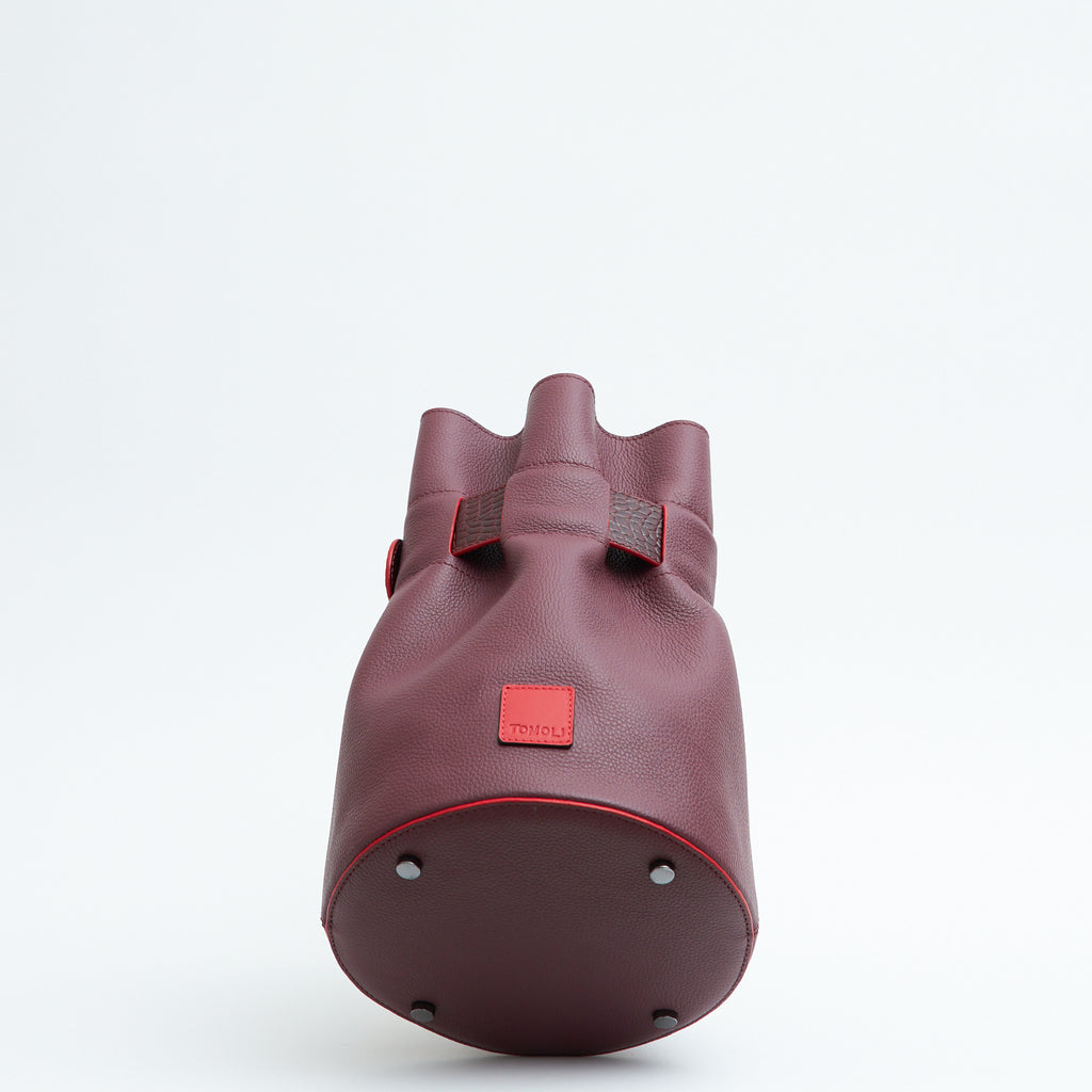 A product photo showing the bottom perspective view of a leather bucket bag. The bag is burgundy red and has a bold belt cinch in croc-embossed leather. The TOMOLI logo is debossed on a small red square towards the bottom of the bag. The bottom is round and has four gunmetal rivets.