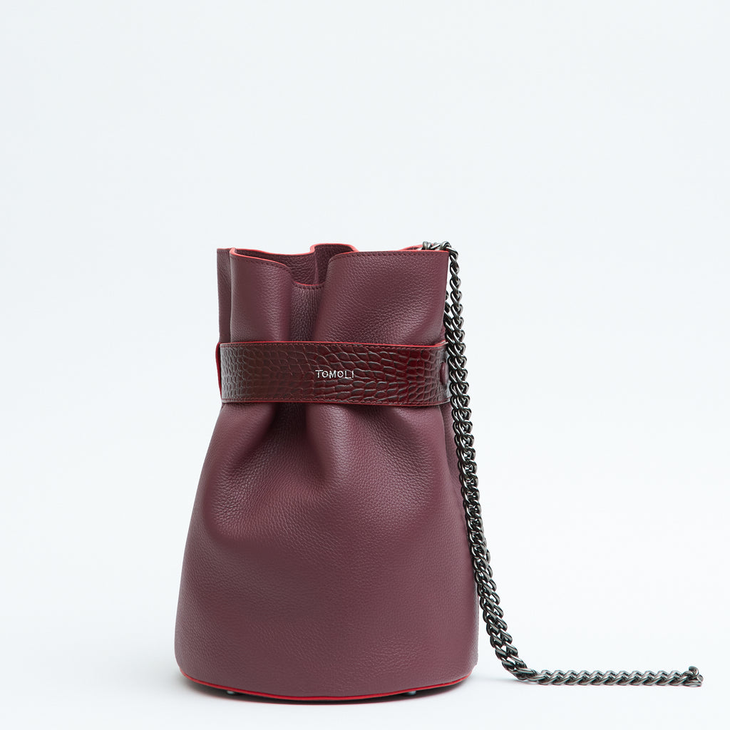 The product photo of a fashion handbag showing a burgundy red leather bucket bag with a croc-embossed belt cinch and a chain strap. This is the front view of the Tomoli La Bourse Mini leather bucket bag in Sangria.