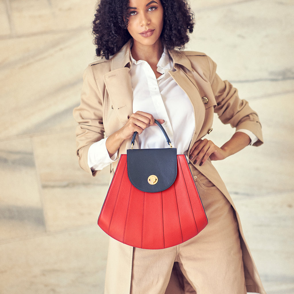 A fashion and style editorial photo showing a model wearing a neutral outfit and holding a bright red leather handbag. The handbag has a shell-like shape and is color-blocked with a dark blue flap closure and handle. The bag adds a pop of color to the neutral background. This is the Tomoli Gisel handbag in Shady Red.