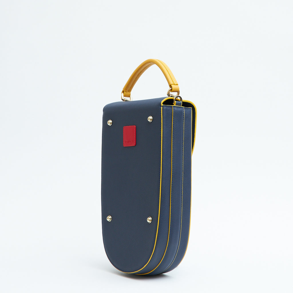 A fashion product photo showing the back view of a colorful blue leather handbag. The bag has an elongated saddle shape with a yellow top handle, and yellow trims. There is a small red square and gold metal rivets. This is the Tomoli Fitini II structured saddle handbag in Golden Denim. This handbag can be used as an everyday crossbody bag, a work bag, or a special occasion bag.