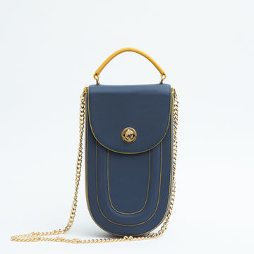 A fashion product photo showing the front view of a colorful blue leather handbag. The bag has an elongated saddle shape with a flap closure, a yellow top handle, and yellow trims. There is a chain strap and a metal lock in gold hardware. This is the Tomoli Fitini II structured saddle handbag in Golden Denim. This handbag can be used as an everyday crossbody bag, a work bag, or a special occasion bag.