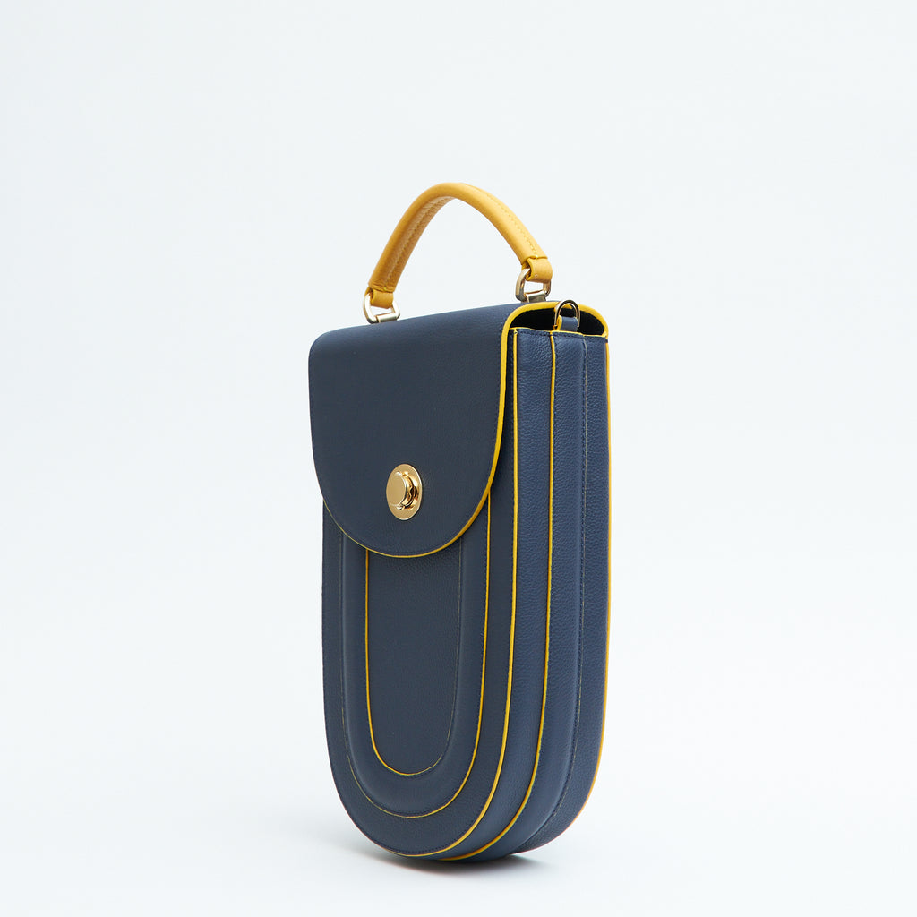 A fashion product photo showing the side view of a colorful blue leather handbag. The bag has an elongated saddle shape with a flap closure, a yellow top handle, and yellow trims. There is a chain strap and a metal lock in gold hardware. This is the Tomoli Fitini II structured saddle handbag in Golden Denim. This handbag can be used as an everyday crossbody bag, a work bag, or a special occasion bag.
