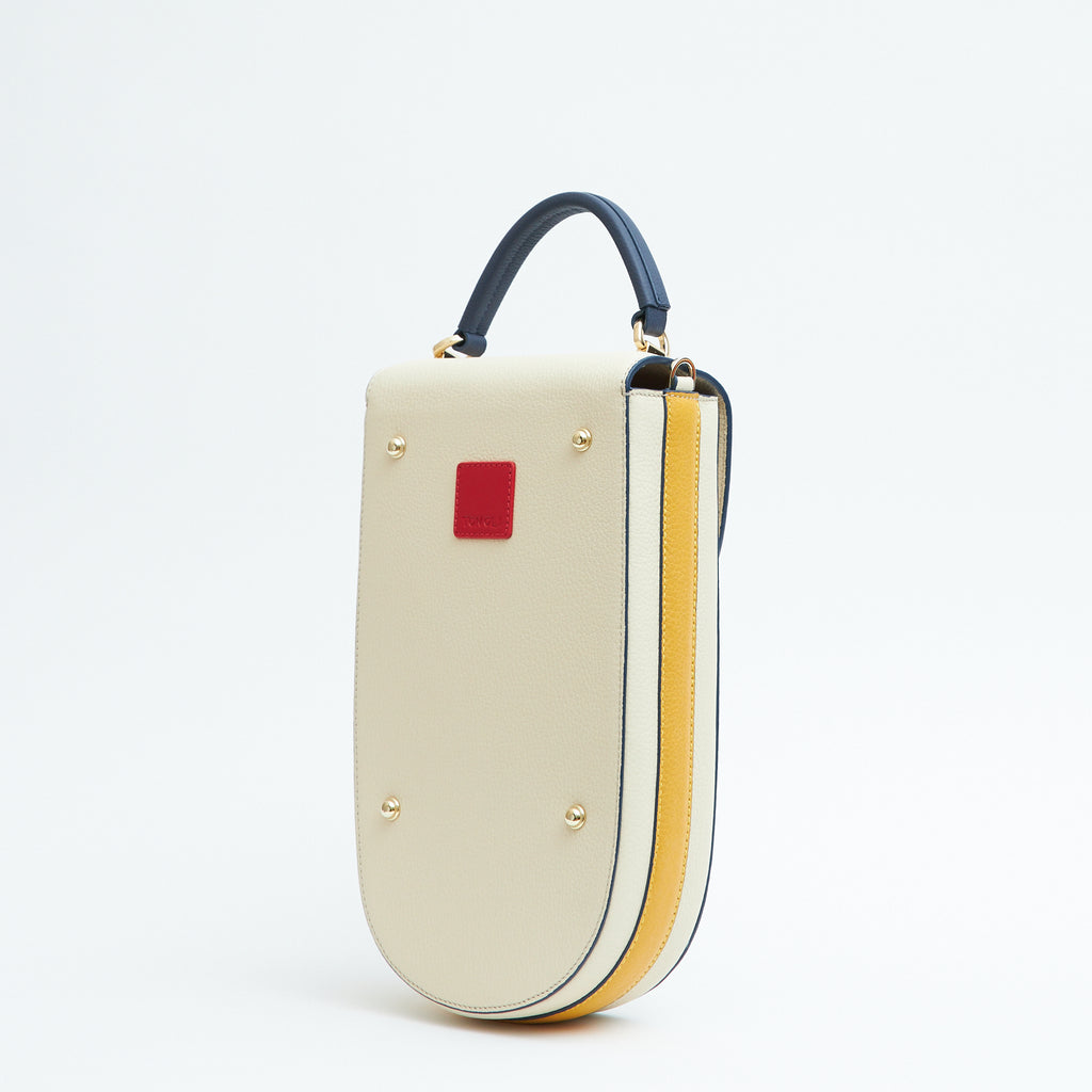 A fashion product photo showing the back view of a color-blocked off-white leather handbag. The bag has an elongated rounded shape that resembles a saddle bag. The handbag has dark blue trims, a dark blue handle, and a yellow stripe on the side. There is a small red square and gold metal rivets. This is the Tomoli Fitini II structured saddle handbag in Eclectic Ivory. This handbag can be used as an everyday crossbody bag, a work bag, or a special occasion bag.