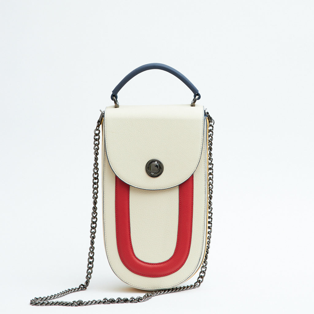 A fashion product photo showing the front view of a color-blocked off-white, blue and red leather handbag. The bag has an elongated rounded shape that resembles a saddle bag. The handbag has a flap closure with dark blue trims and a dark blue handle. There is a chain strap and a metal lock in gunmetal hardware. This is the Tomoli Fitini II structured saddle handbag in Eclectic Ivory. This handbag can be used as an everyday crossbody bag, a work bag, or a special occasion bag.