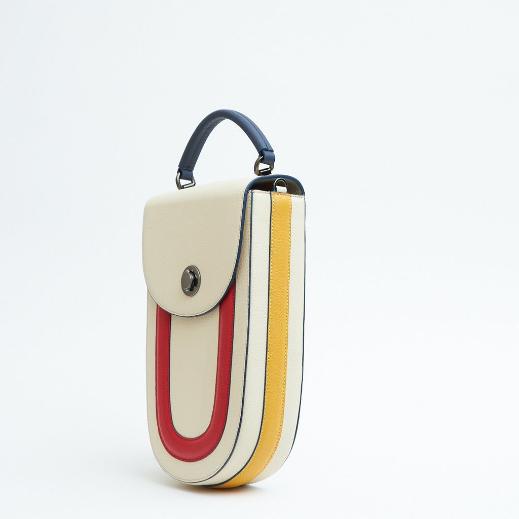 A product photo showing the side view of a color-blocked off-white leather handbag. The bag has an elongated rounded shape that resembles a saddle bag. The handbag has a flap closure with dark blue trims, a dark blue handle, and red and yellow stripes. There is a chain strap and a metal lock in gunmetal hardware. This is the Tomoli Fitini II structured saddle handbag in Eclectic Ivory. This handbag can be used as an everyday crossbody bag, a work bag, or a special occasion bag.