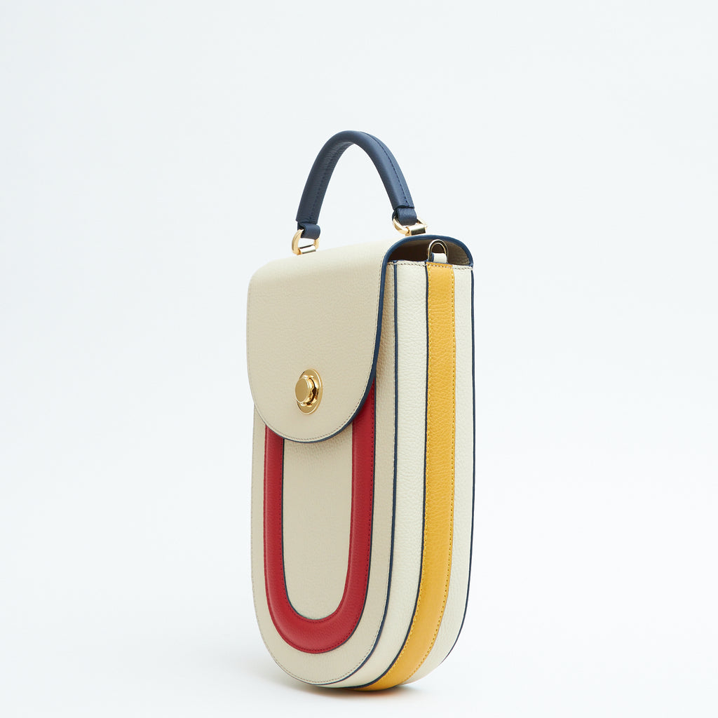 A product photo showing the side view of a color-blocked off-white leather handbag. The bag has an elongated rounded shape that resembles a saddle bag. The handbag has a flap closure with dark blue trims, a dark blue handle, and red and yellow stripes. There is a chain strap and a metal lock in gold hardware. This is the Tomoli Fitini II structured saddle handbag in Eclectic Ivory. This handbag can be used as an everyday crossbody bag, a work bag, or a special occasion bag.
