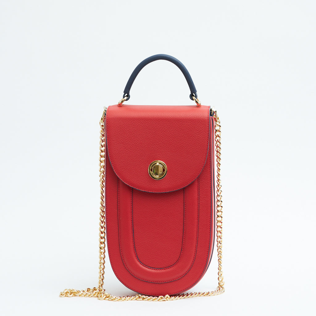 A fashion product photo showing the front view of a colorful red leather handbag. The bag has an elongated saddle shape with a flap closure, a dark blue top handle, and dark blue trims. There is a chain strap and a metal lock in gold hardware. This is the Tomoli Fitini II structured saddle handbag in Shady Red. This handbag can be used as an everyday crossbody bag, a work bag, or a special occasion bag.