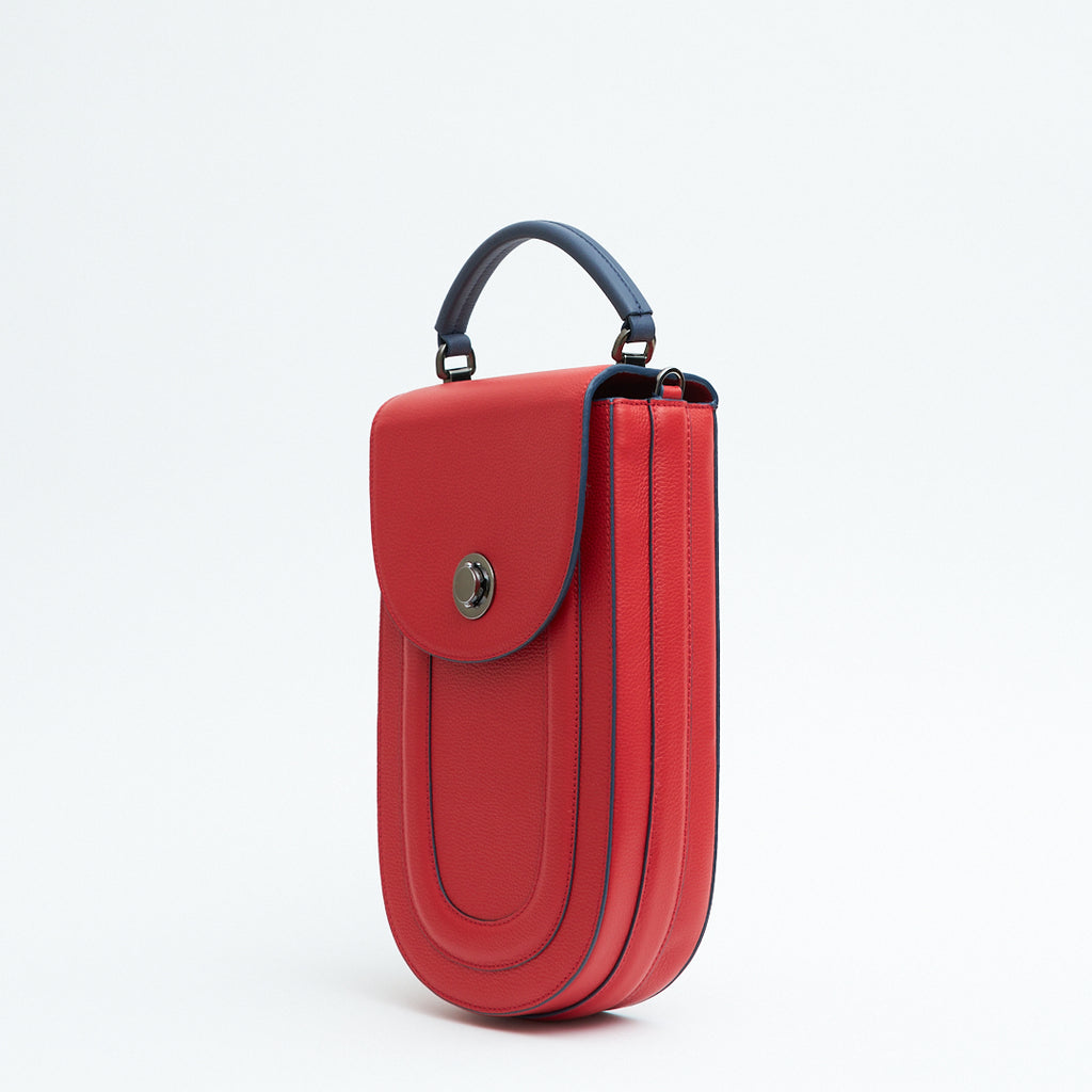 A fashion product photo showing the side view of a colorful red leather handbag. The bag has an elongated saddle shape with a flap closure, a dark blue top handle, and dark blue trims. There is a chain strap and a metal lock in gunmetal hardware. This is the Tomoli Fitini II structured saddle handbag in Shady Red. This handbag can be used as an everyday crossbody bag, a work bag, or a special occasion bag.