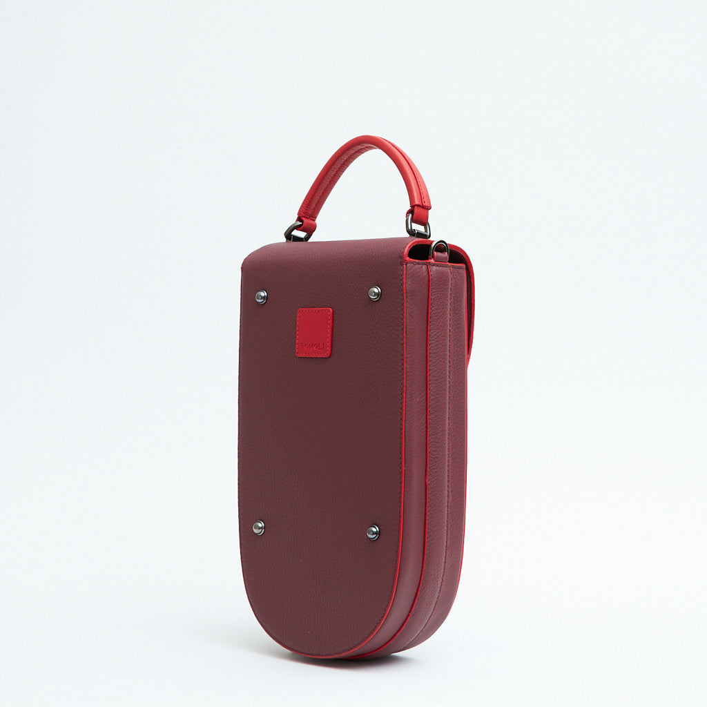 A fashion product photo showing the back view of a colorful burgundy red leather handbag. The bag has an elongated saddle shape with a red top handle, and red trims. There is a small red square and gunmetal metal rivets. This is the Tomoli Fitini II structured saddle handbag in Hot Sangria. This handbag can be used as an everyday crossbody bag, a work bag, or a special occasion bag.