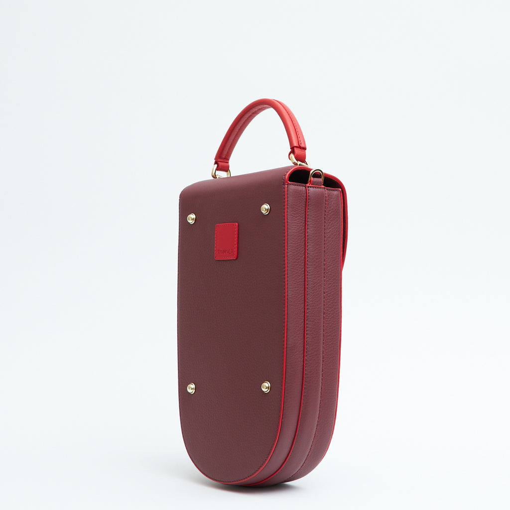 A fashion product photo showing the back view of a colorful burgundy red leather handbag. The bag has an elongated saddle shape with a red top handle, and red trims. There is a small red square and gold metal rivets. This is the Tomoli Fitini II structured saddle handbag in Hot Sangria. This handbag can be used as an everyday crossbody bag, a work bag, or a special occasion bag.