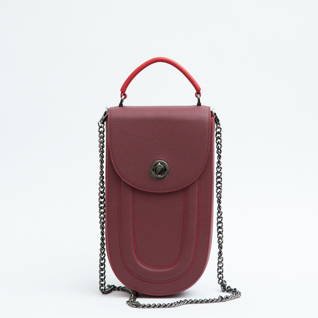 A fashion product photo showing the front view of a burgundy red leather handbag. The bag has an elongated saddle shape with a flap closure, a red top handle, and red trims. There is a chain strap and a metal lock in gunmetal hardware. This is the Tomoli Fitini II structured saddle handbag in Hot Sangria. This handbag can be used as an everyday crossbody bag, a work bag, or a special occasion bag.