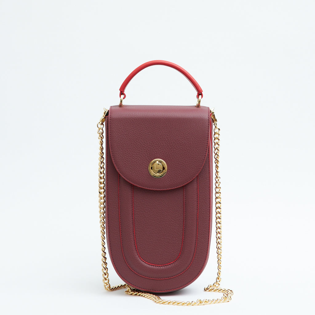 A fashion product photo showing the front view of a colorful burgundy red leather handbag. The bag has an elongated saddle shape with a flap closure, a red top handle, and red trims. There is a chain strap and a metal lock in gold hardware. This is the Tomoli Fitini II structured saddle handbag in Hot Sangria. This handbag can be used as an everyday crossbody bag, a work bag, or a special occasion bag.