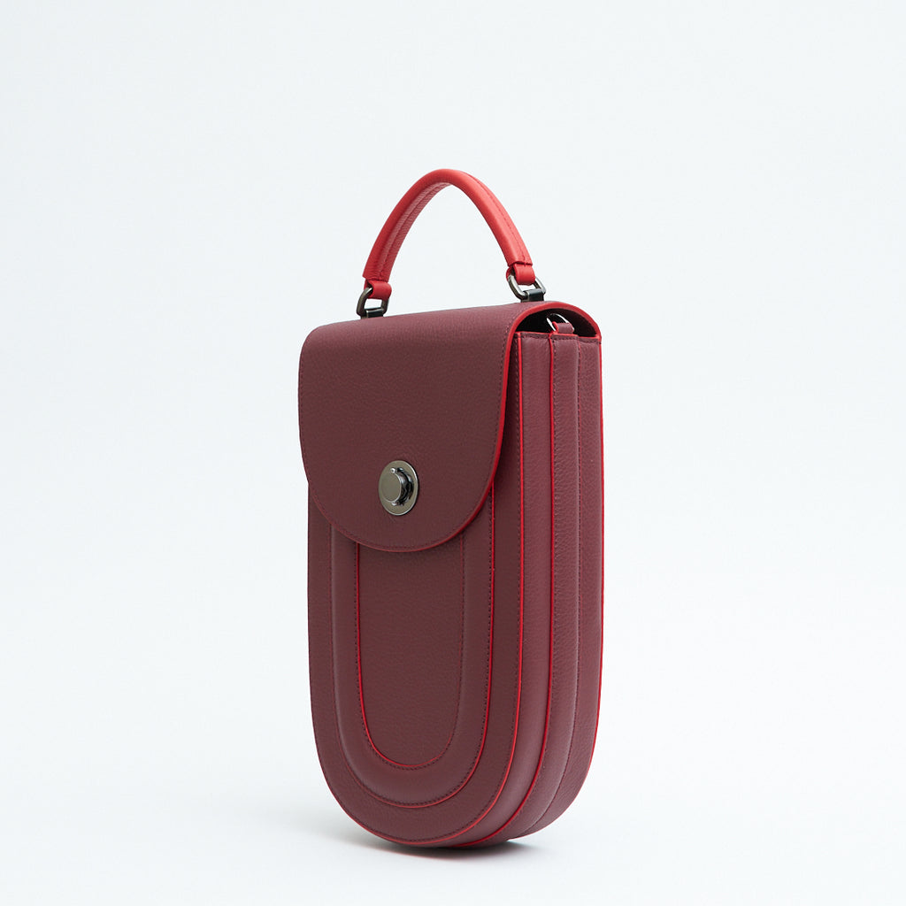 A fashion product photo showing the side view of a burgundy red leather handbag. The bag has an elongated saddle shape with a flap closure, a red top handle, and red trims. There is a chain strap and a metal lock in gunmetal hardware. This is the Tomoli Fitini II structured saddle handbag in Hot Sangria. This handbag can be used as an everyday crossbody bag, a work bag, or a special occasion bag.