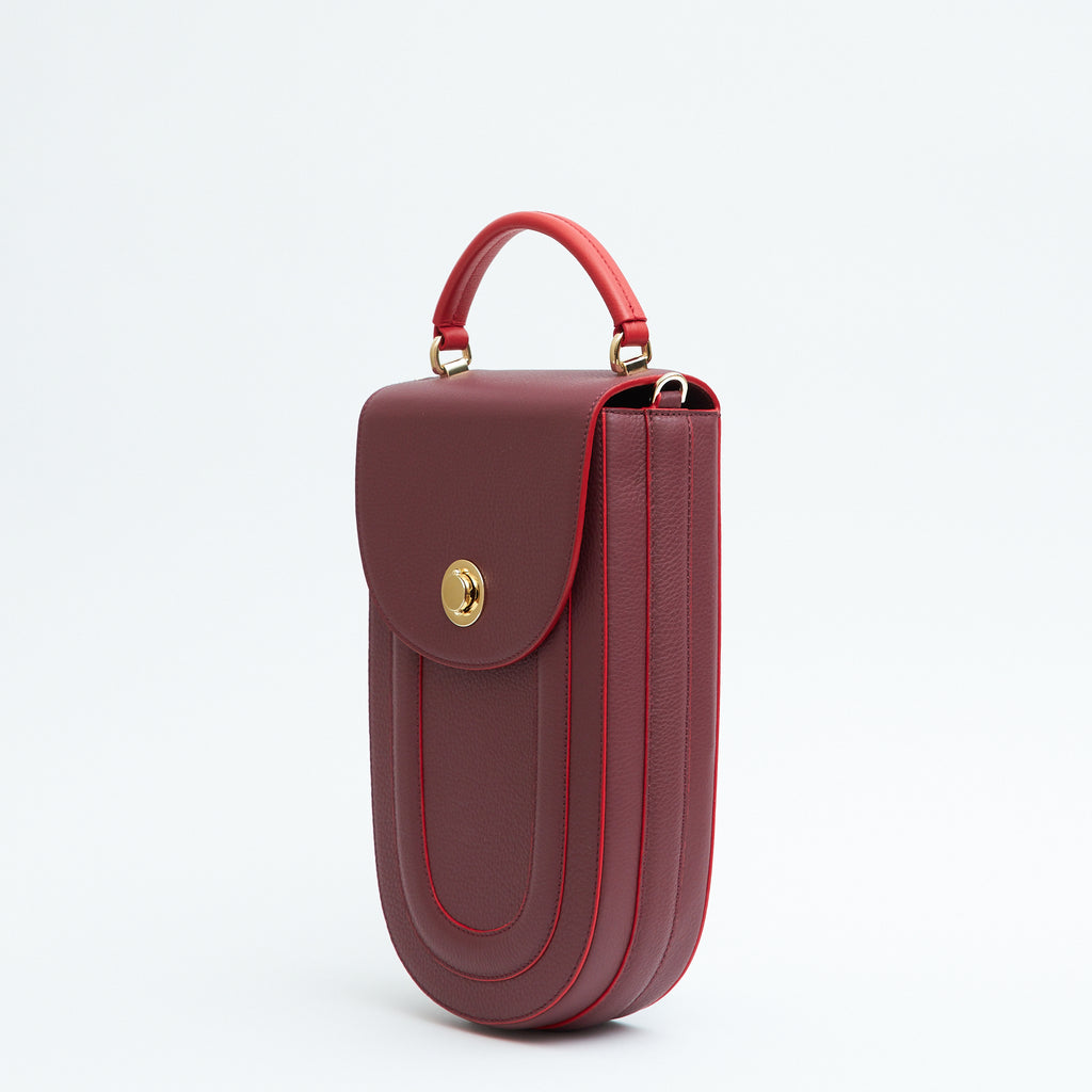 A fashion product photo showing the side view of a colorful burgundy red leather handbag. The bag has an elongated saddle shape with a flap closure, a red top handle, and red trims. There is a chain strap and a metal lock in gold hardware. This is the Tomoli Fitini II structured saddle handbag in Hot Sangria. This handbag can be used as an everyday crossbody bag, a work bag, or a special occasion bag.