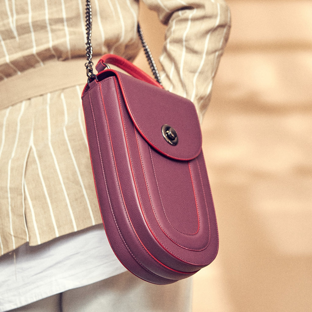 A fashion and style editorial photo showing a model wearing a neutral outfit with a burgundy red leather handbag. The bag has an elongated saddle shape with a flap closure, a red top handle, and red trims. There is a chain strap and a metal lock in gunmetal hardware. This is the Tomoli Fitini II structured saddle handbag in Hot Sangria. This handbag can be used as an everyday crossbody bag, a work bag, or a special occasion bag.
