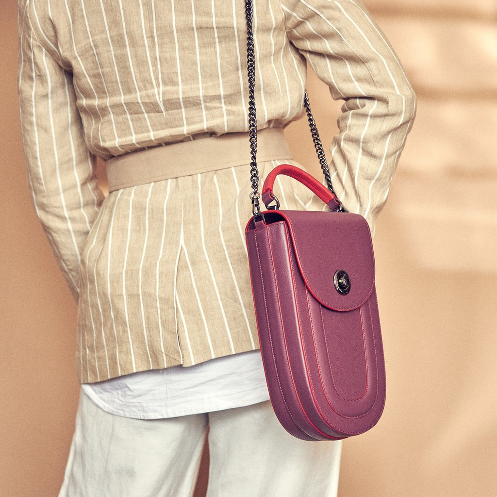 A fashion and style editorial photo showing a model wearing a neutral outfit with a burgundy red leather handbag. The bag has an elongated saddle shape with a flap closure, a red top handle, and red trims. There is a chain strap and a metal lock in gunmetal hardware. This is the Tomoli Fitini II structured saddle handbag in Hot Sangria. This handbag can be used as an everyday crossbody bag, a work bag, or a special occasion bag.