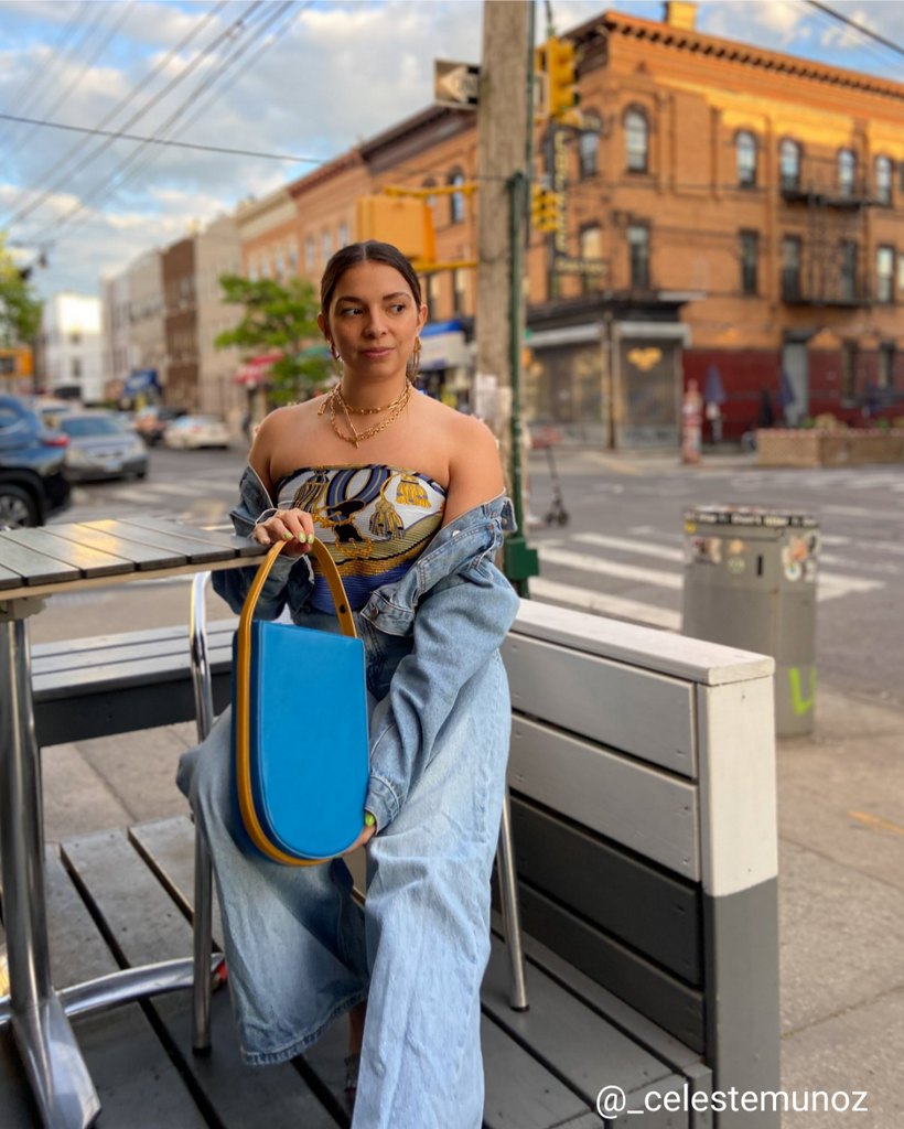 A fashion and style photo showing influencer @_celestemunoz wearing a denim outfit with a color-blocked blue and yellow leather bag. The bag has a distinctive rounded shape and a contouring handle. The influencer is wearing blue jeans with a blue denim jacket. The overall outfit appears colorful on a neutral city background. The bag is the Tomoli Fitini in Golden Sky.