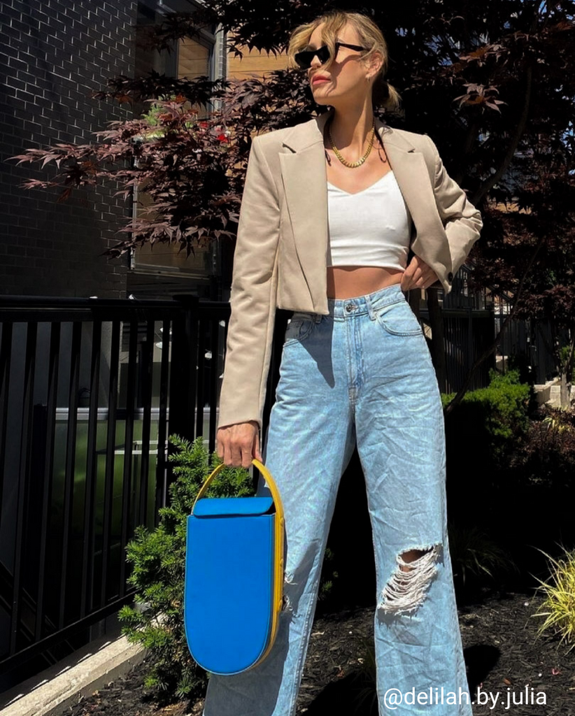 A fashion and style photo showing influencer @_delilah.by.julia wearing a denim outfit with a color-blocked blue and yellow leather bag. The bag has a distinctive rounded shape and a contouring handle. The influencer is wearing blue jeans with a cropped top and a jacket. The bag adds a pop of color to the neutral outfit. The bag is the Tomoli Fitini in Golden Sky.