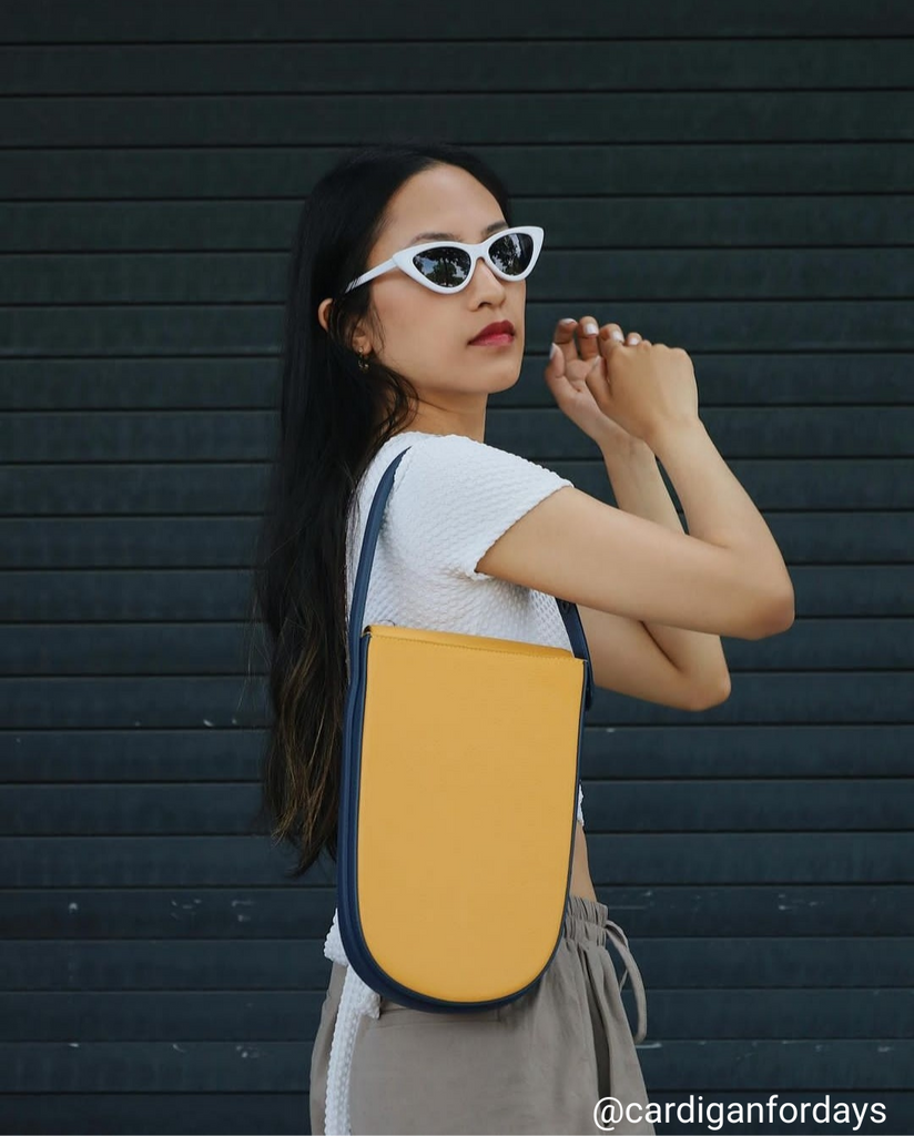 A fashion and style photo showing influencer @cardiganfordays wearing a summer outfit with a yellow leather bag. The bag has a distinctive rounded shape and is color-blocked with a dark blue handle. The woman is wearing bold sunglasses that have a white frame.