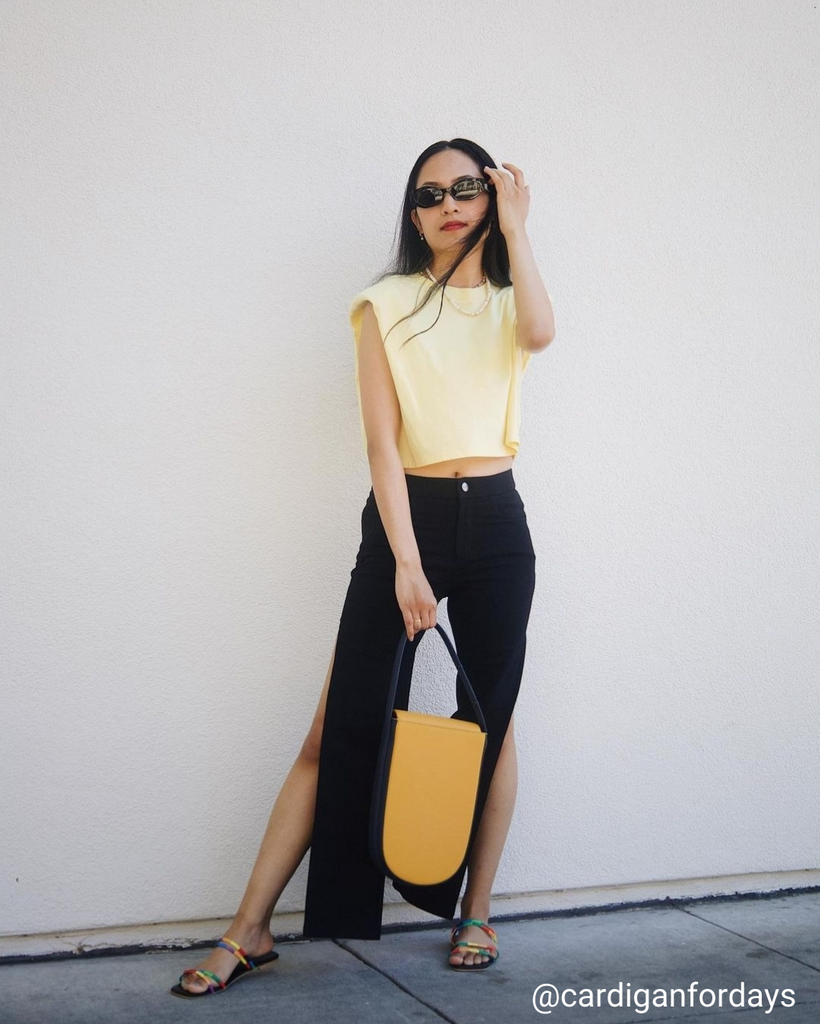 A fashion and style photo showing influencer @cardiganfordays wearing a summer outfit with a yellow bag. The bag has a distinctive rounded shape and a contrasting blue handle. The influencer is wearing black pants with slits and a cropped yellow top.