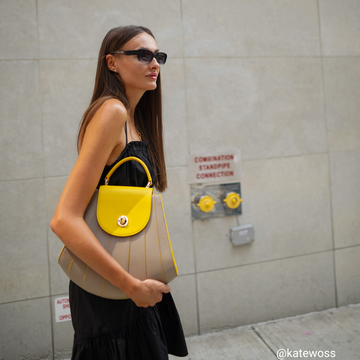 A fashion and style editorial photo showing influencer @katewoss wearing the Tomoli Gisel handbag in Sun Stone. The bag is color-blocked with yellow and beige and has a shell-like shape with a rounded flap closure.