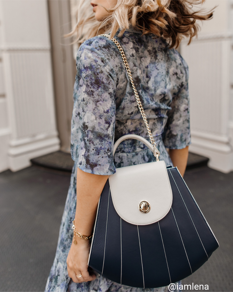 A fashion and style editorial photo showing style influencer @iamlena wearing a floral dress with a color-blocked blue and white leather handbag. The bag has a seashell shape with stripes and a rounded flap closure. The influencer is wearing the blue shoulder bag using a chain strap.