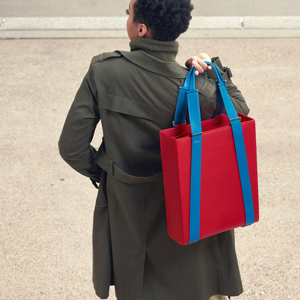 An outfit and style inspiration photo on how to wear a large tote bag. A woman wearing a green trench coat is holding a large red leather tote bag. The handbag has a structured rectangular briefcase shape and is color-blocked with blue straps. This is the Tomoli Kora large convertible leather tote in Sky Red.