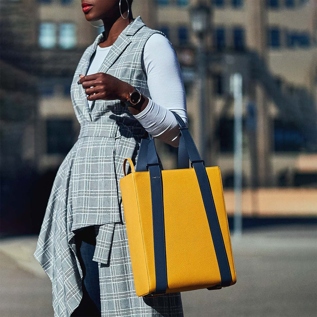 An outfit and style inspiration photo on how to wear a large tote bag. A woman wearing a plaid sleeveless coat is holding a large yellow leather tote handbag. The bag has a structured rectangular briefcase shape and is color-blocked with dark blue straps. This is the Tomoli Kora large convertible leather tote in Shady Maize.