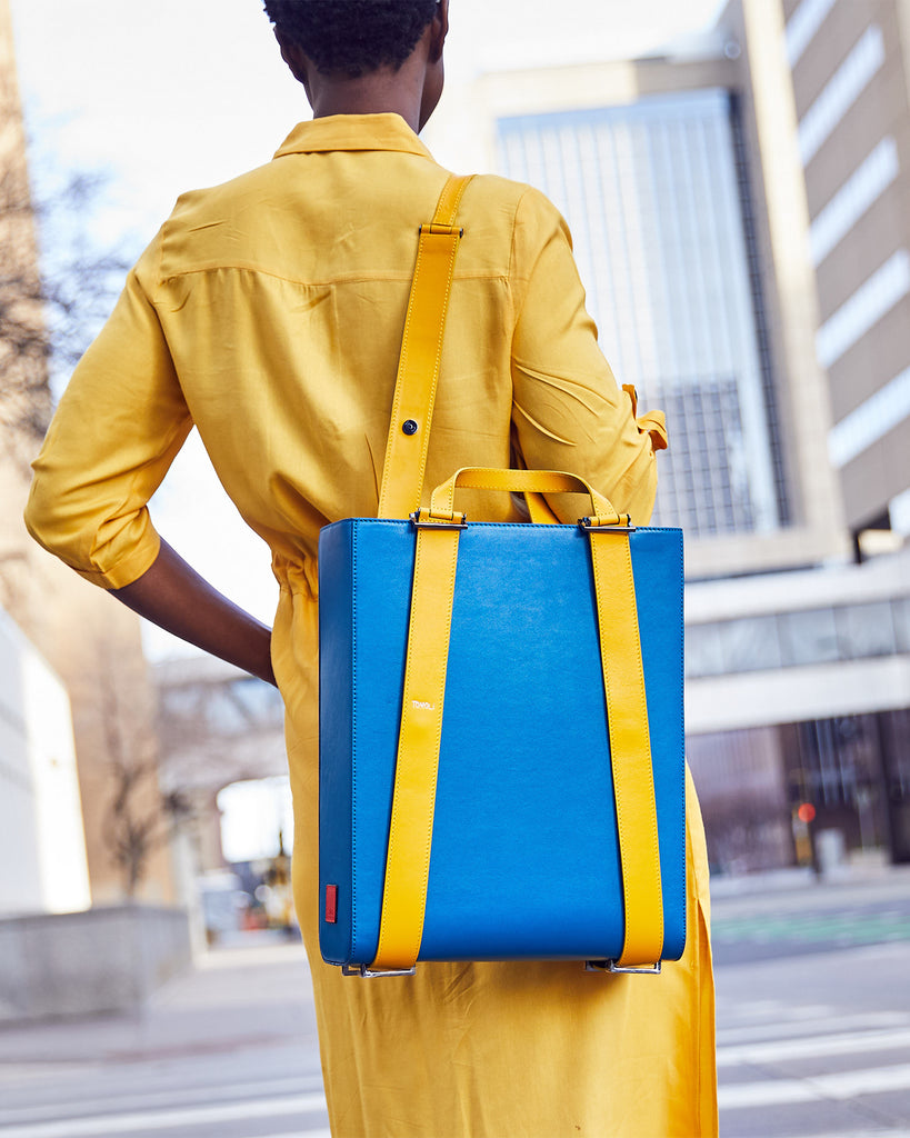 A fashion and style editorial photo showing a woman wearing a teal leather tote bag. The tote bag has a structured rectangular briefcase shape and color-blocked yellow leather straps. This is the Tomoli Kora large convertible leather tote in golden teal. The bag can fit a laptop and can be styled for work or to elevate casual outfits.