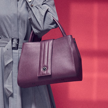 A fashion and style photo showing a model holding a burgundy red leather tote handbag. The handbag has a classic trapeze shape and a quilted flap closure. This is the Tomoli Briffani Lean interchangeable tote handbag in Sangria. This bag can be used as an everyday bag, a work tote, or an oversized clutch.