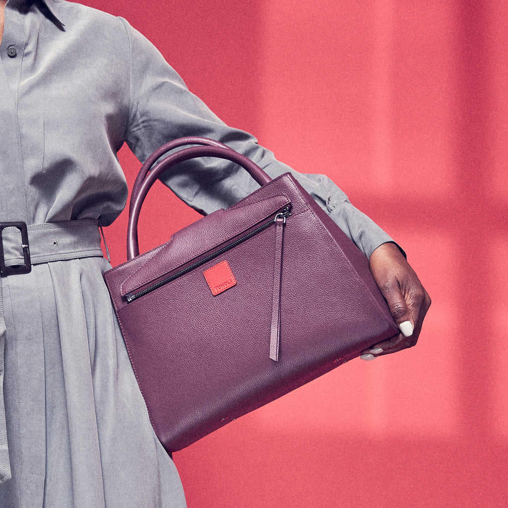 A fashion and style photo showing a model holding a burgundy red leather handbag. The handbag has a classic trapeze shape with a back pocket, a zipper to interchange flaps, and top handles. This is the back view of the Tomoli Briffani Lean interchangeable tote handbag in Sangria. This bag can be used as an everyday bag, a work tote, or even an oversized clutch.