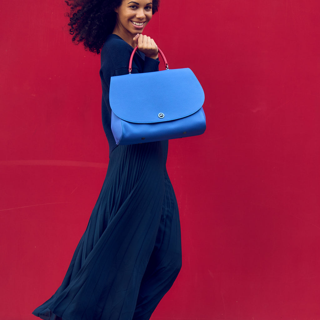 The photo of a woman showing a colorful blue purple and red top handle leather handbag. The handbag has a blue purple oversized round flap cover on a blue purple body. The handle is red. This is the Tomoli Briffani Jut in Hot Iris.