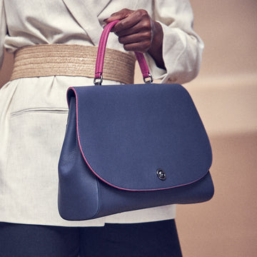 A fashion and style photo showing a model holding a dark blue flap leather handbag. The handbag has an oversized round flap cover that is color-blocked with a pink handle. This is the Tomoli Briffani Jut in Flirty Denim.