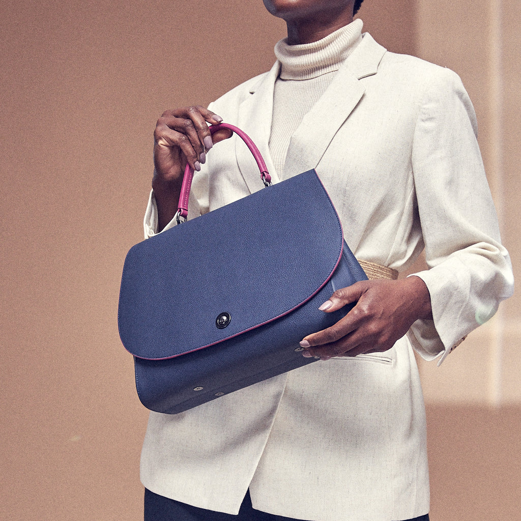 A fashion and style photo showing a model holding a dark blue flap leather handbag. The handbag has an oversized round flap cover that is color-blocked with a pink handle. This is the Tomoli Briffani Jut in Flirty Denim.