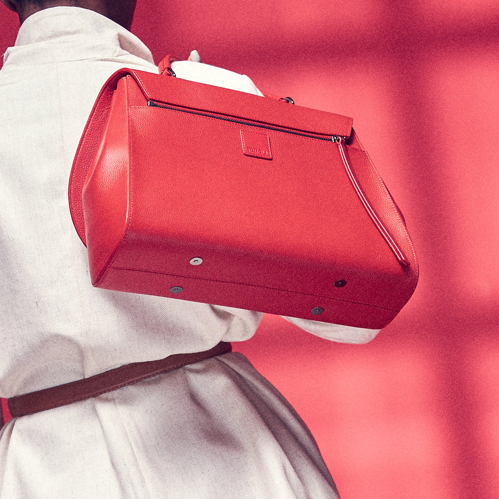 A fashion and style editorial photo showing a model holding a red top handle leather handbag. The bag has a minimalistic design and the back has a zipper attachment. The overall look appears color-blocked with red and white. This is the Tomoli Briffani Jut interchangeable satchel in red.