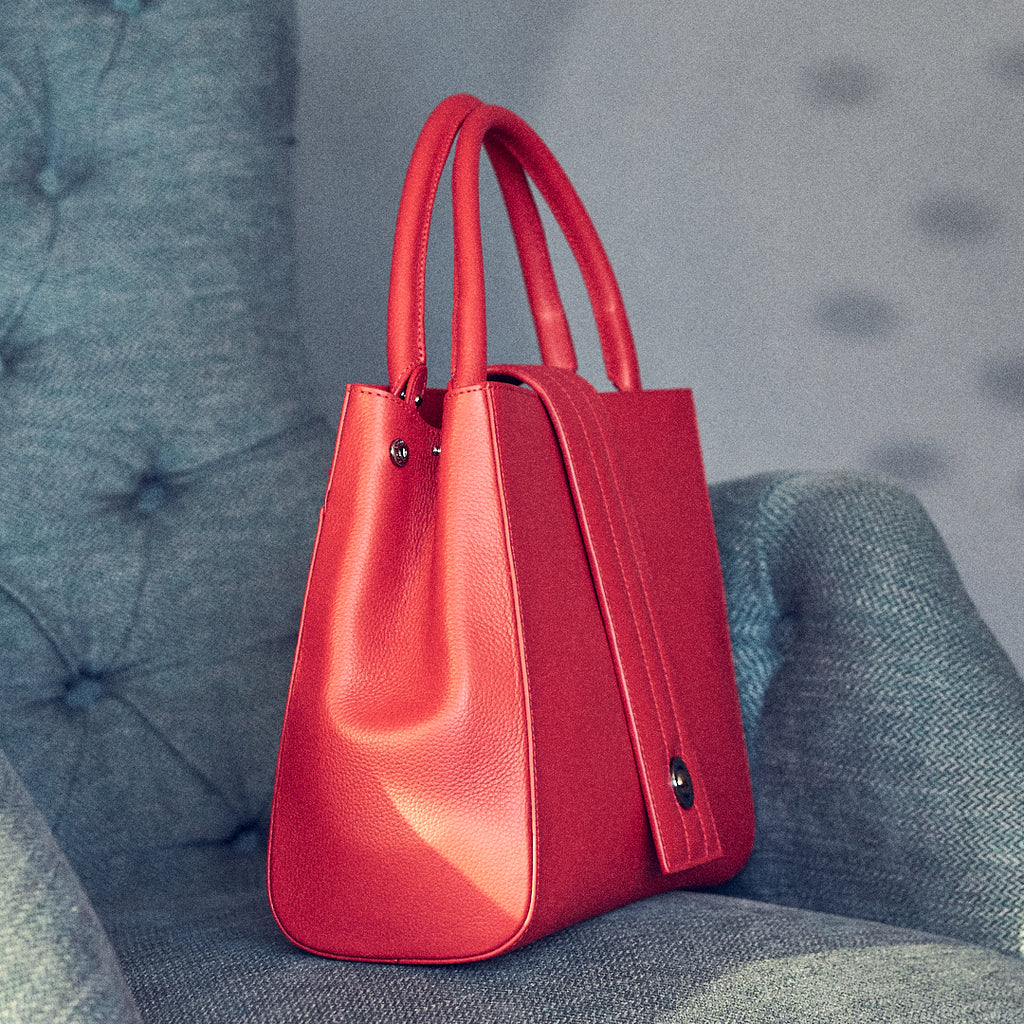 The product photo of a red leather handbag. The handbag has a classic trapeze shape with a straight quilted flap in the middle and top handles. This is the side view of the Tomoli Briffani Lean interchangeable tote handbag in Red. This bag can be used as an everyday bag, a work tote, or even an oversized clutch.