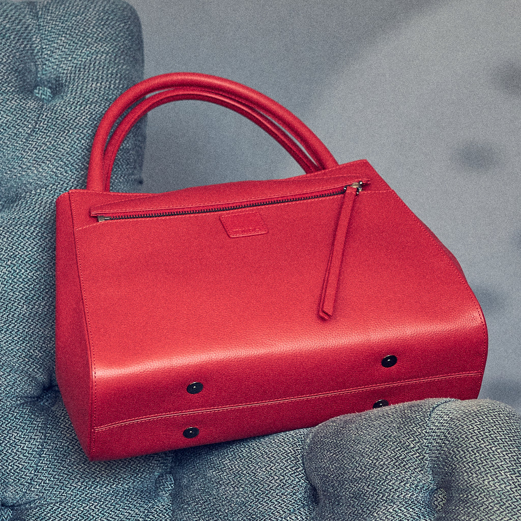 The product photo of a red leather handbag. The handbag has a classic trapeze shape with a back pocket, a zipper to interchange flaps, and top handles. This is the back view of the Tomoli Briffani Lean interchangeable tote handbag in red. This bag can be used as an everyday bag, a work tote, or even an oversized clutch.