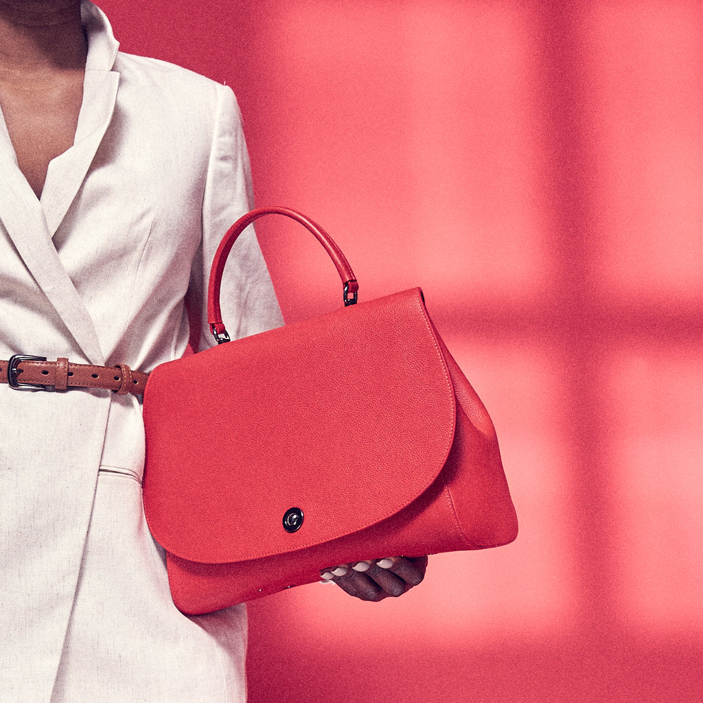 A fashion and style editorial photo showing a model holding a red top handle leather handbag. The bag has a minimalistic design and an oversized rounded flap cover. The overall look appears color-blocked with red and white. This is the Tomoli Briffani Jut interchangeable satchel in red.