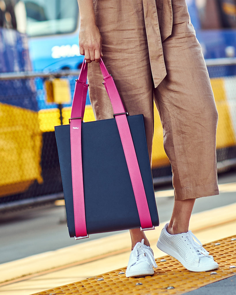An outfit and style inspiration photo on how to wear a large tote bag. A woman wearing a neutral outfit is holding a large dark blue leather tote bag. The handbag has a structured rectangular shape and is color-blocked with pink straps. This is the Tomoli Kora large convertible leather tote in Flirty Denim.